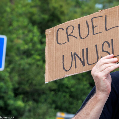 A road sign that says Alligator Alcatraz and an individual in front of it holding a sign that says "Cruel and Unusual."