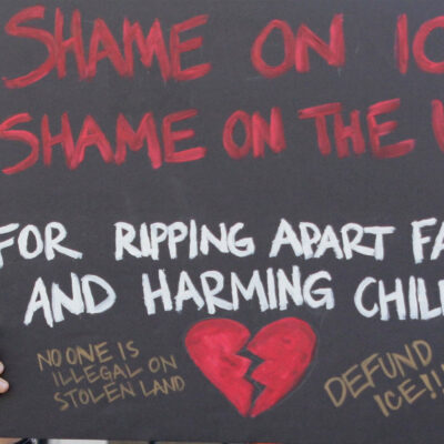 A demonstrator protests ICE and the Trump Administration policy of separating migrant families by holding up a sign (written in red ink on a black background with an illustrated broken red heart at the bottom) that reads, "SHAME ON ICE SHAME ON THE USA FOR RIPPIING APART FAMILIES AND HARMING CHILDREN No One is Illegal On Stolen Land, Defund ICE!!!"