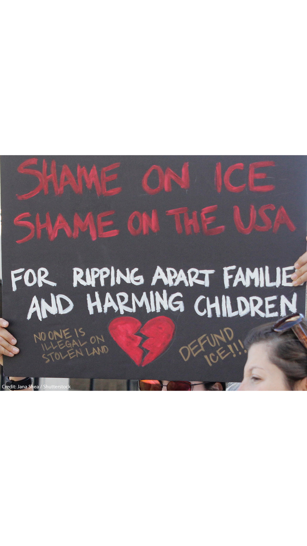 A demonstrator protests ICE and the Trump Administration policy of separating migrant families by holding up a sign (written in red ink on a black background with an illustrated broken red heart at the bottom) that reads, "SHAME ON ICE SHAME ON THE USA FOR RIPPIING APART FAMILIES AND HARMING CHILDREN No One is Illegal On Stolen Land, Defund ICE!!!"