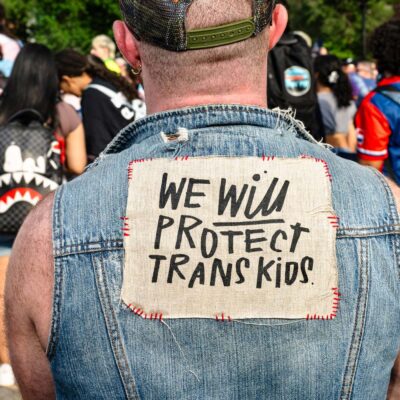 At a trans rally, person (wearing a denim vest and with their back to the camera) displays the sign stitched to the vest that reads "WE WILL PROTECT TRANS KIDS."