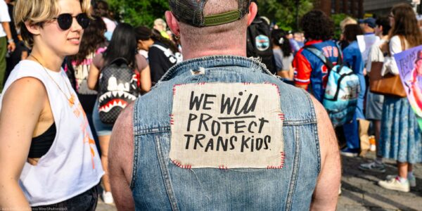 At a trans rally, person (wearing a denim vest and with their back to the camera) displays the sign stitched to the vest that reads "WE WILL PROTECT TRANS KIDS."